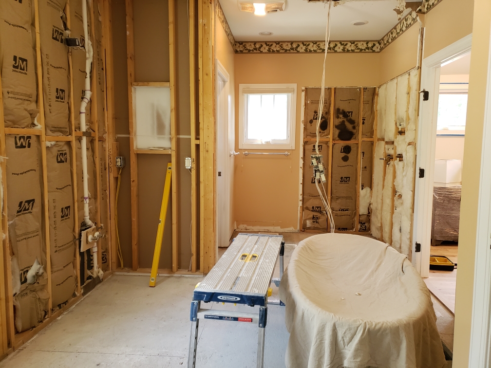 A room under renovation with exposed wall studs and insulation, a covered bathtub, a step ladder, and construction materials scattered around.