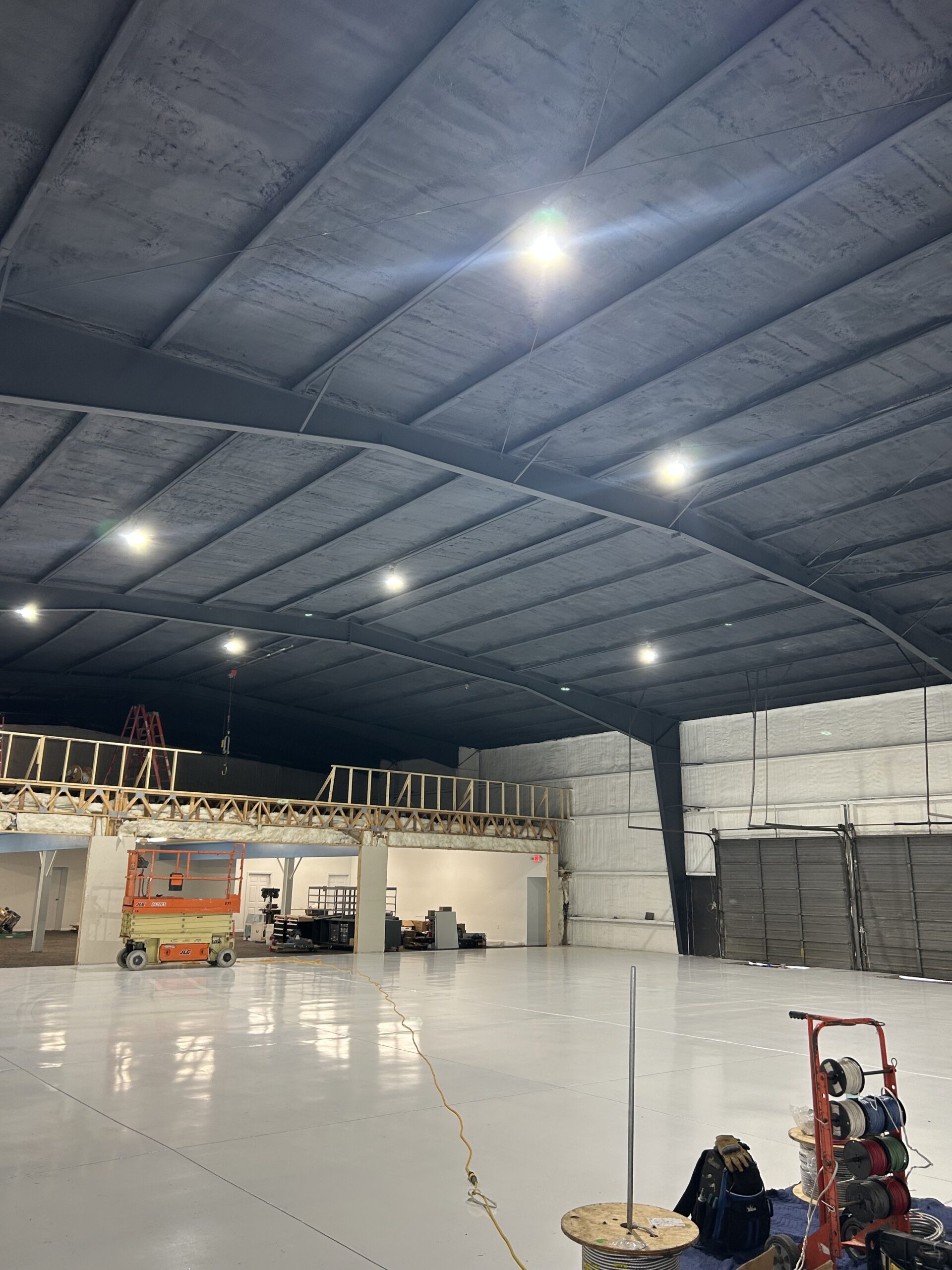 Large industrial warehouse with high curved ceiling, bright overhead lights, construction equipment, and unfinished wooden framing along one wall. Floor is clean and reflective.
