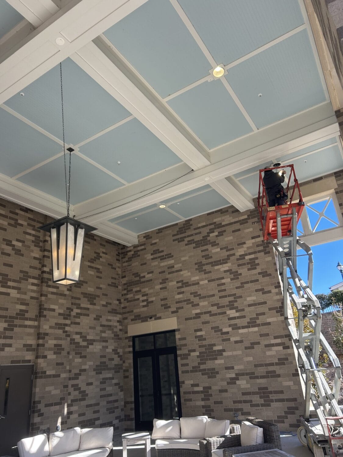 A worker on a red scissor lift is near the ceiling of a covered outdoor patio area with brick walls, light blue paneling, and patio furniture.