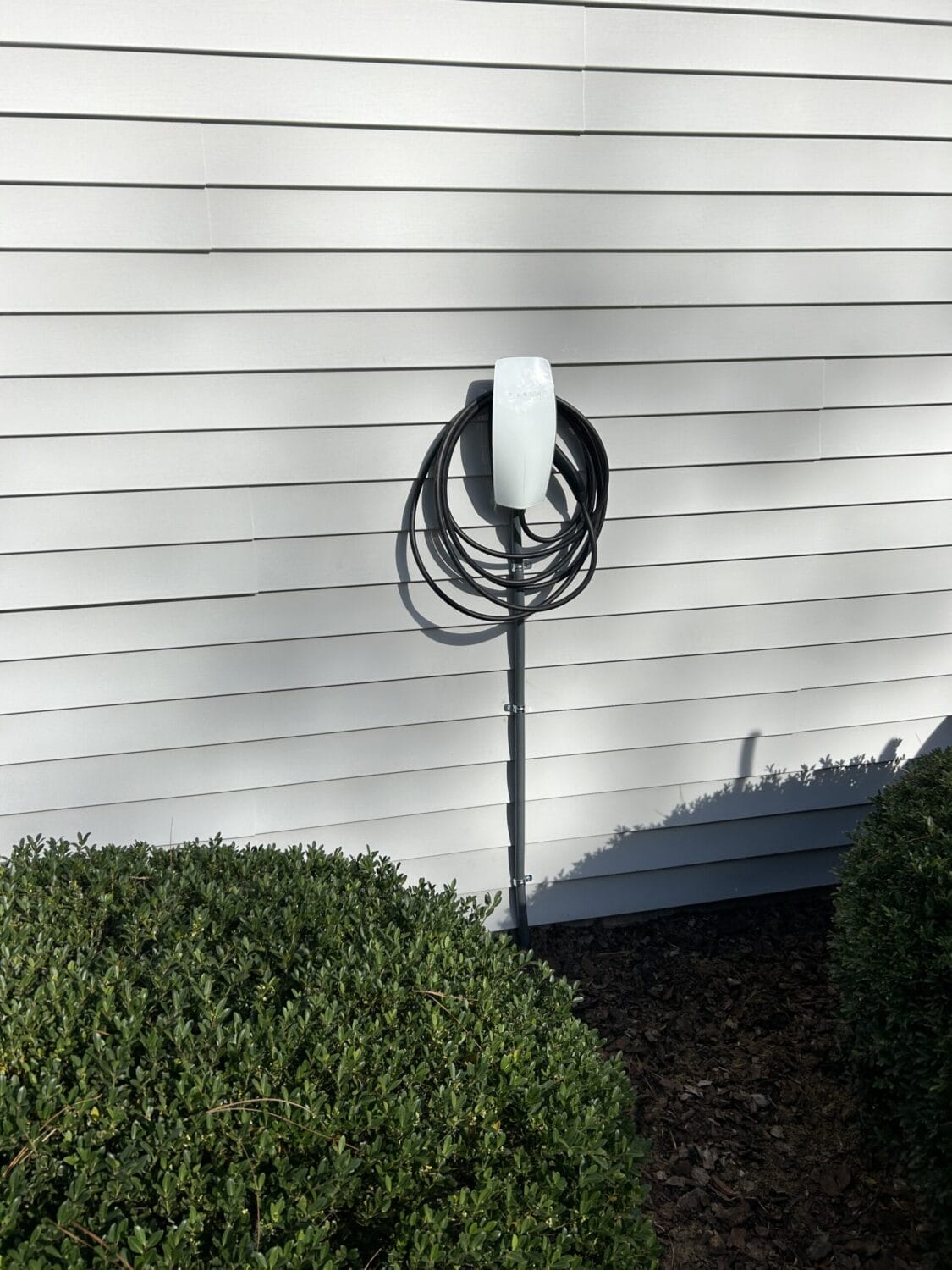 Electric vehicle charging station mounted on an exterior wall with the charging cable neatly coiled, next to some green shrubs.