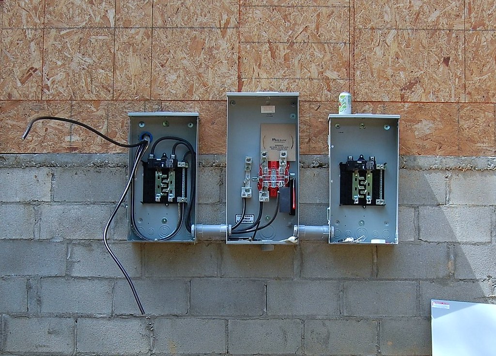 Three open electrical panels mounted on a concrete block wall, showing exposed wires and components, with a beverage can placed on the rightmost panel.