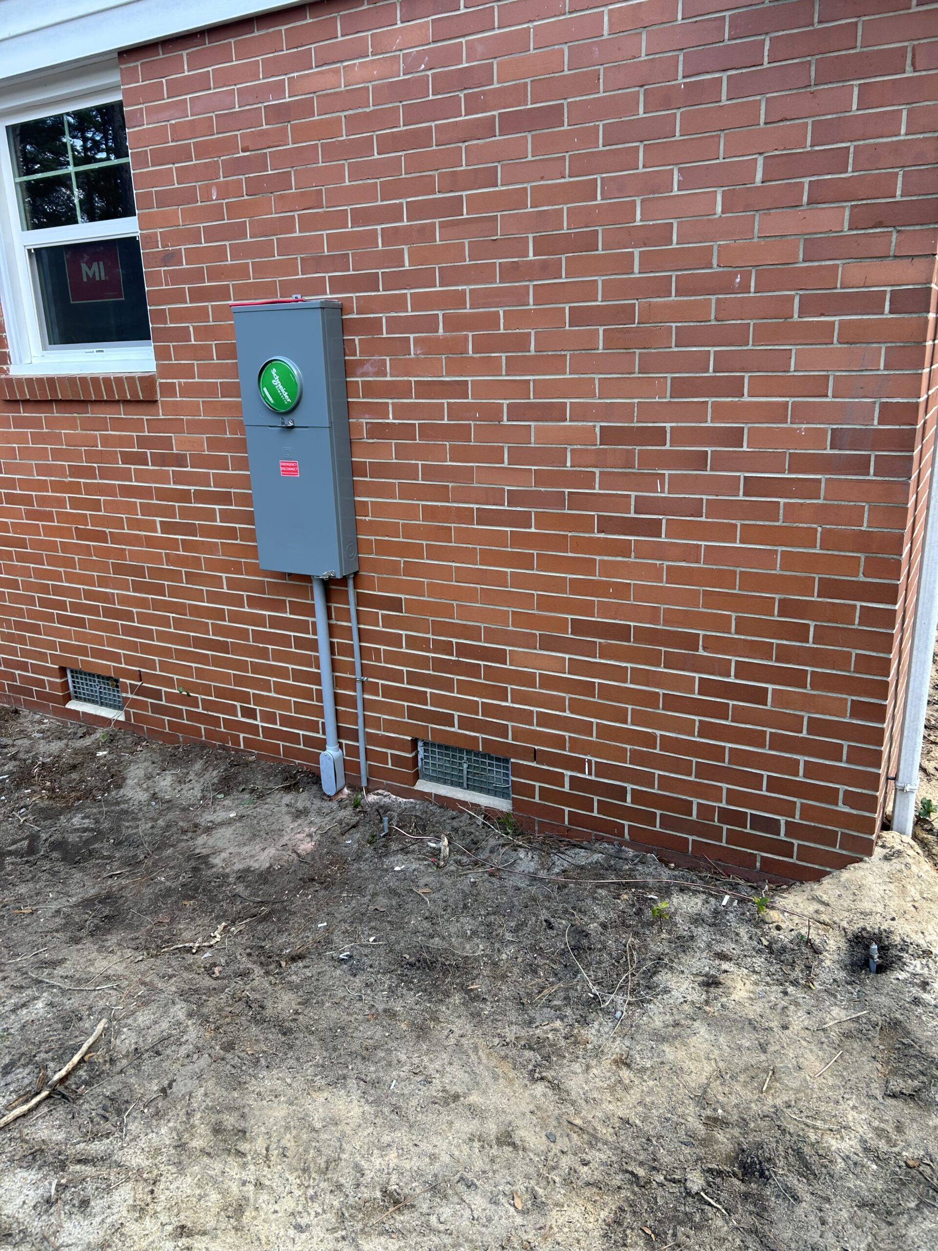 A gray electrical box with a green circular indicator is mounted on a red brick wall next to two ventilation grates and a window. The ground below is bare soil.