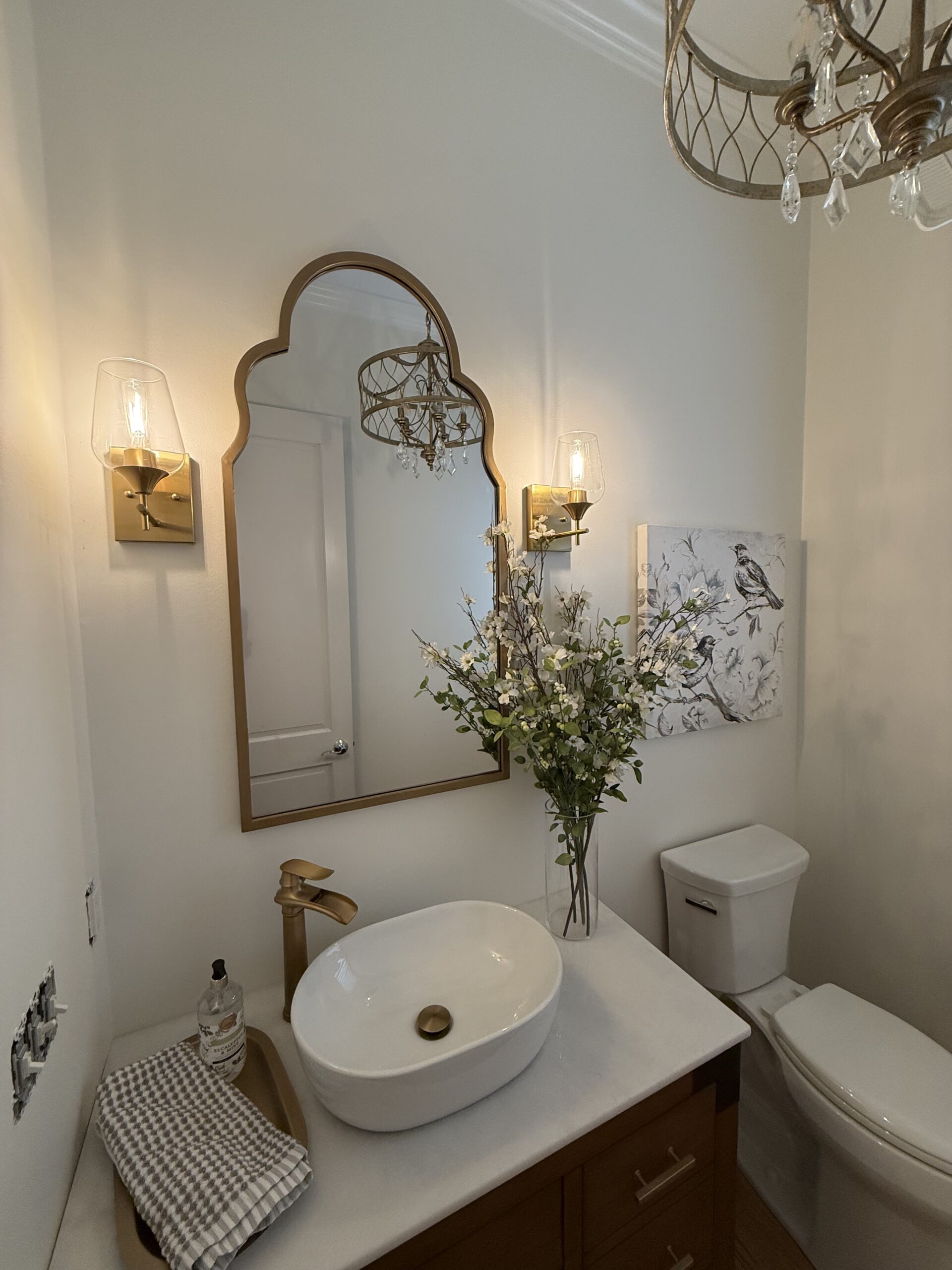 Modern bathroom with a white vessel sink, gold fixtures, mirror, wall art with a bird, fresh flowers in a vase, and a towel on the counter. A chandelier and two wall sconces provide lighting.