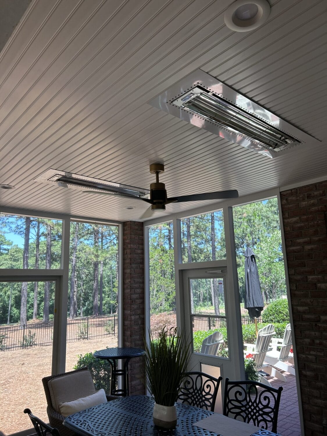 Ceiling with a fan and two rectangular heaters above an outdoor patio area, furnished with a table, chairs, and surrounded by windows overlooking a yard with trees.