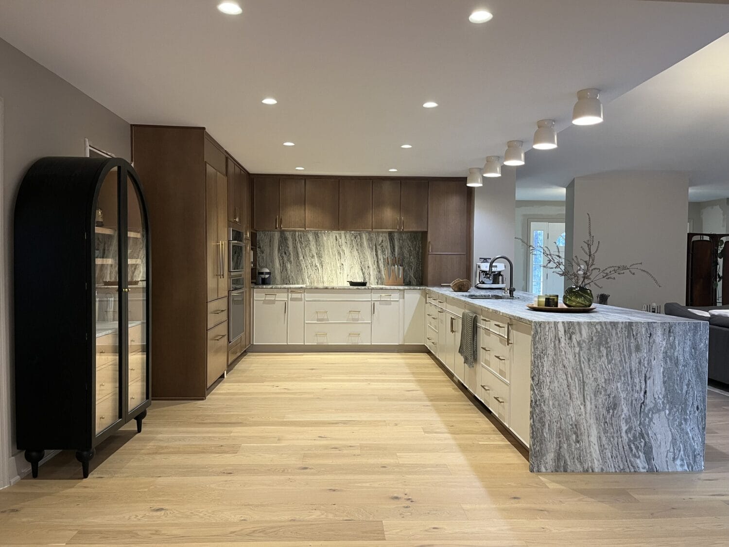 Modern kitchen with wood cabinets, marble backsplash and island, light wood floors, glass cabinet on the left, and spotlights on the ceiling installed by a professional electrician.
