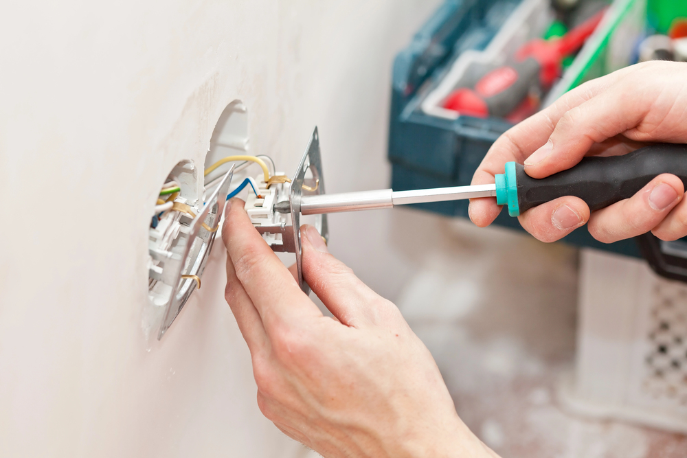 A person installs or repairs an electrical outlet using a screwdriver, with exposed wires visible and a toolbox in the background.