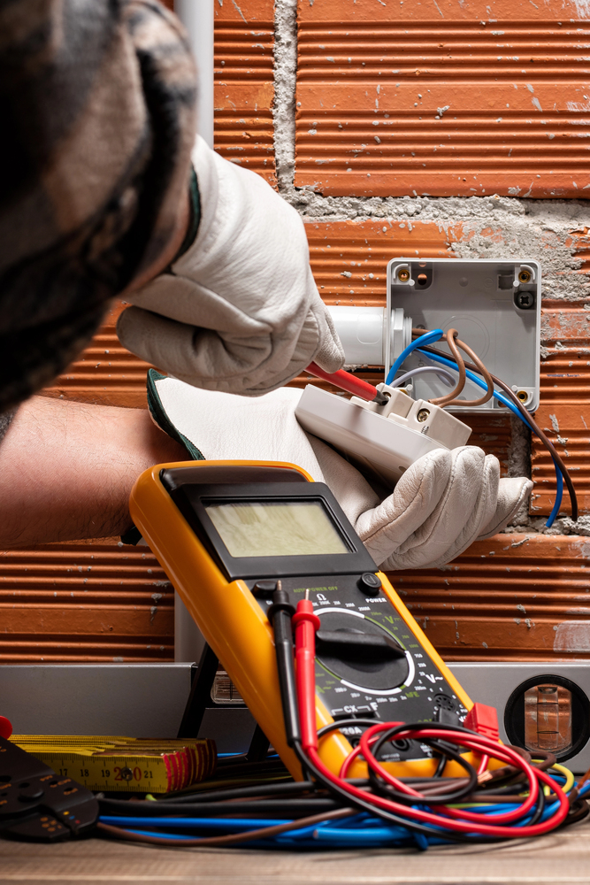 Electrician wearing gloves installs wiring into an electrical outlet box on a brick wall, with a digital multimeter and tools visible in the foreground.