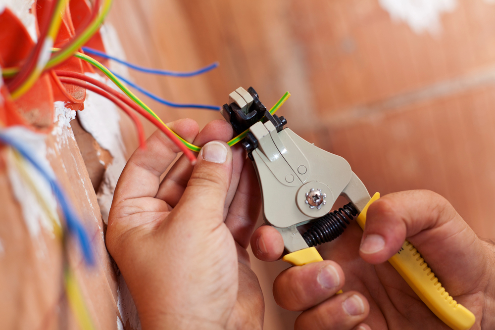A person uses a wire stripping tool to strip the insulation off colored electrical wires near a wall under construction.
