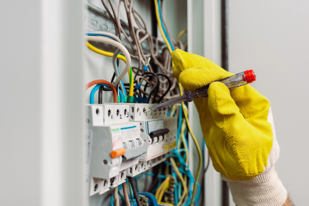A person wearing yellow gloves uses a voltage tester on wiring inside an electrical panel with multiple wires and circuit breakers.
