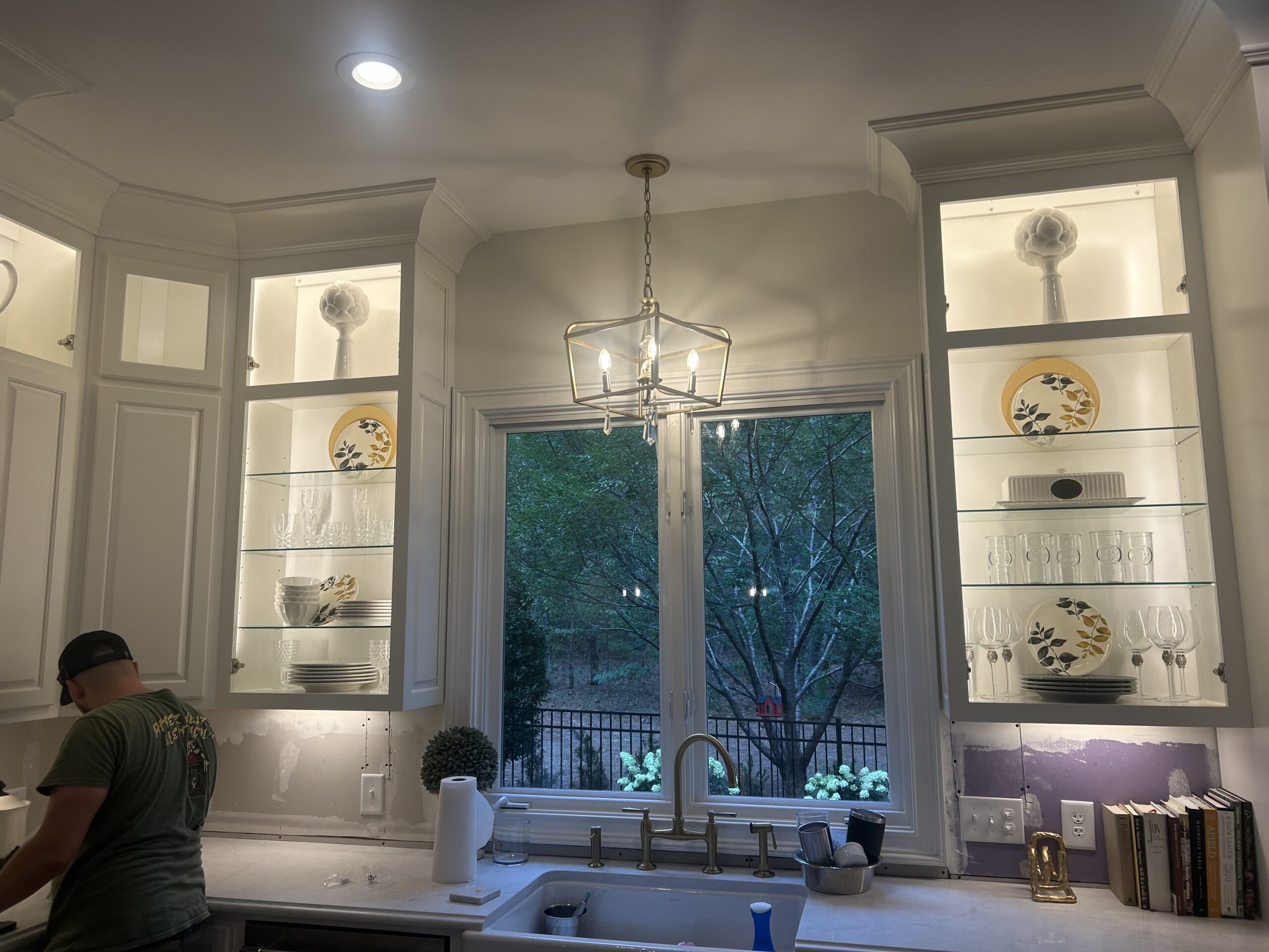 A kitchen sink area with glass shelves displaying plates and glasses, a window overlooking trees, and a person standing to the left, partially out of frame beneath a modern lighting installation.