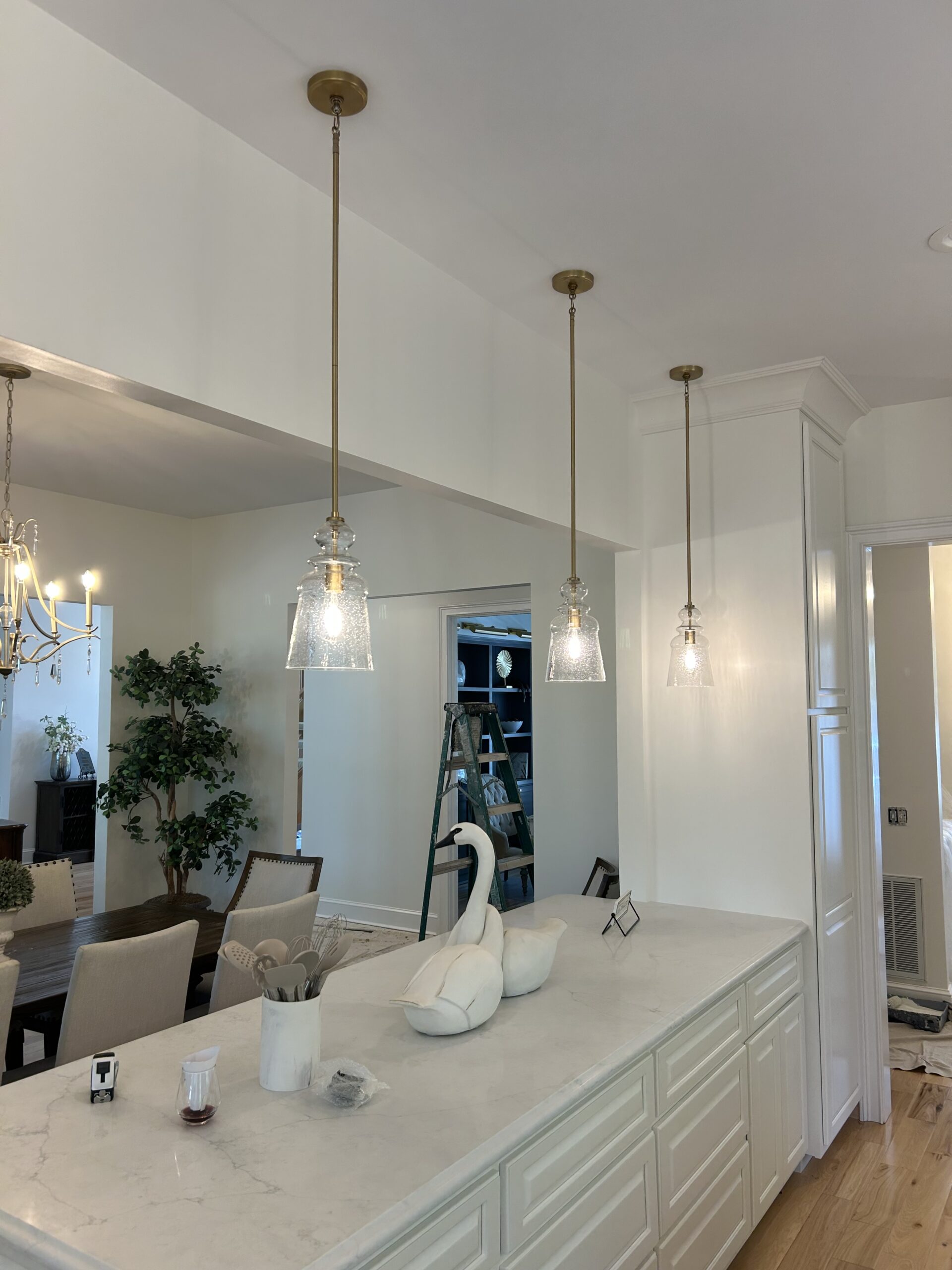 A kitchen island with a white countertop features three pendant lights, a white swan decoration, and a dining area in the background. A stepladder and indoor plant are visible.