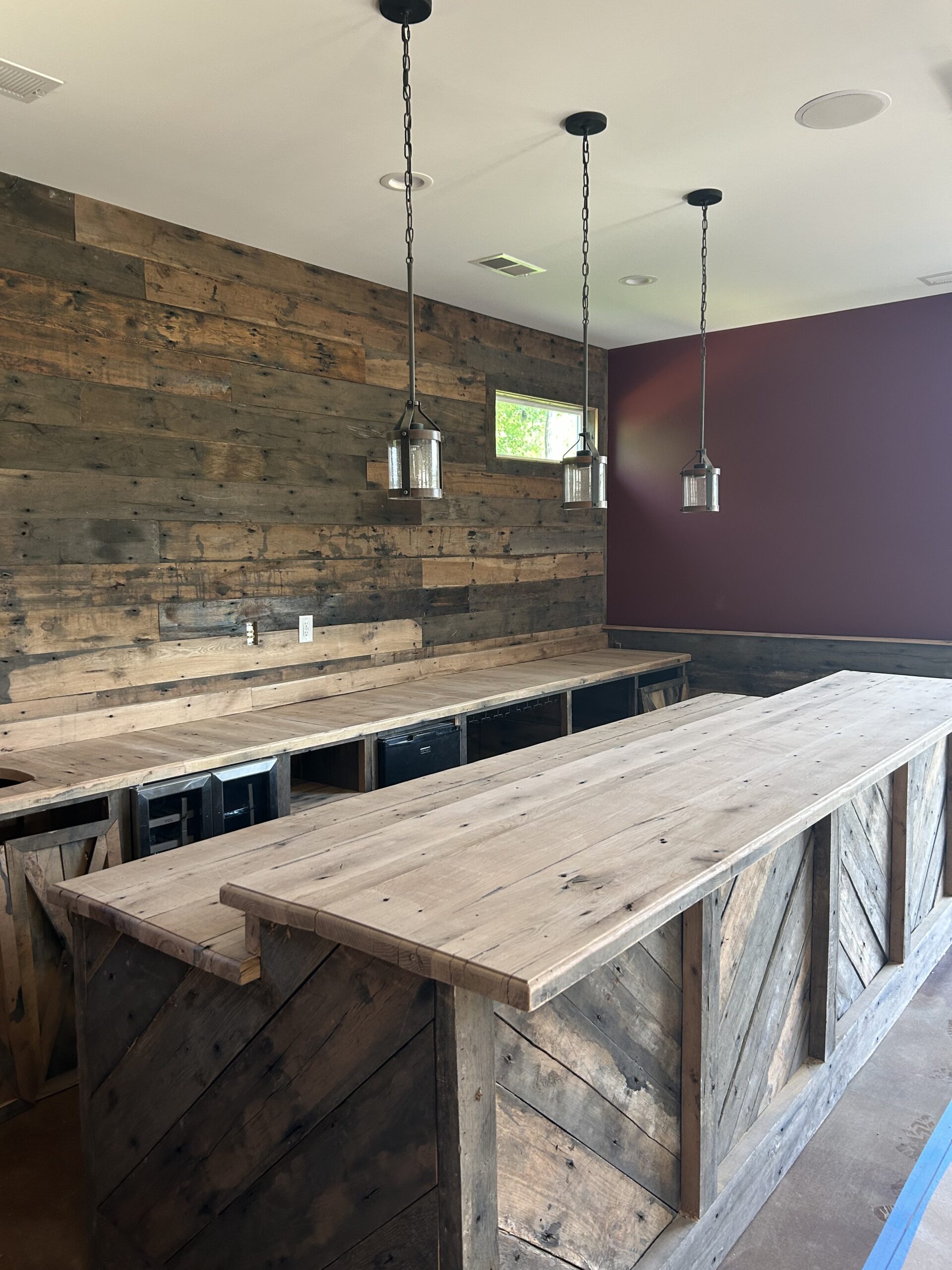 A rustic bar with wooden counters and walls, three pendant lights from a stylish lighting installation hanging above, and a small window letting in natural light.