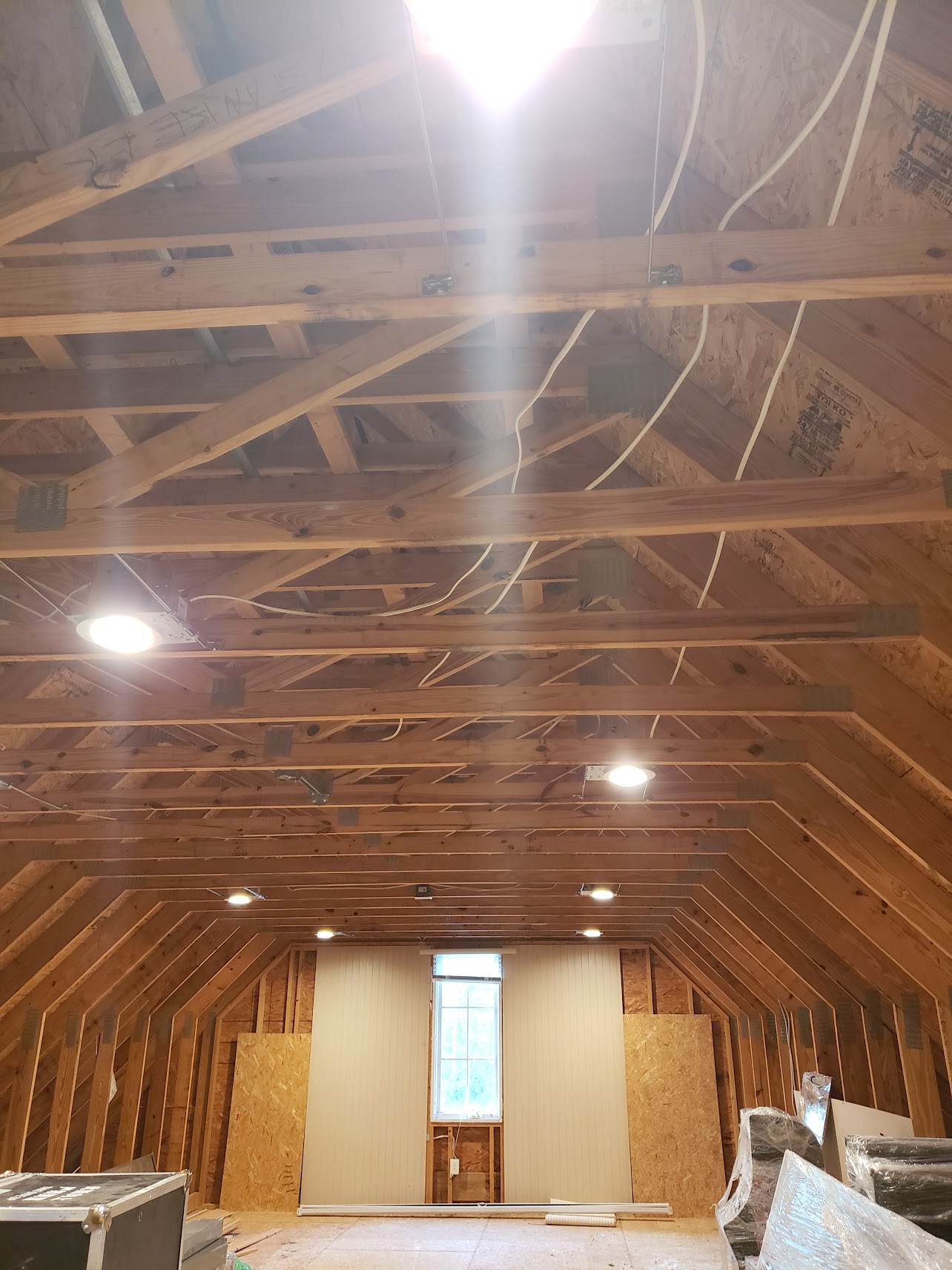 Unfinished attic space with exposed wooden beams, visible wiring, and construction materials on the floor, lit by ceiling lights and natural light from a window.