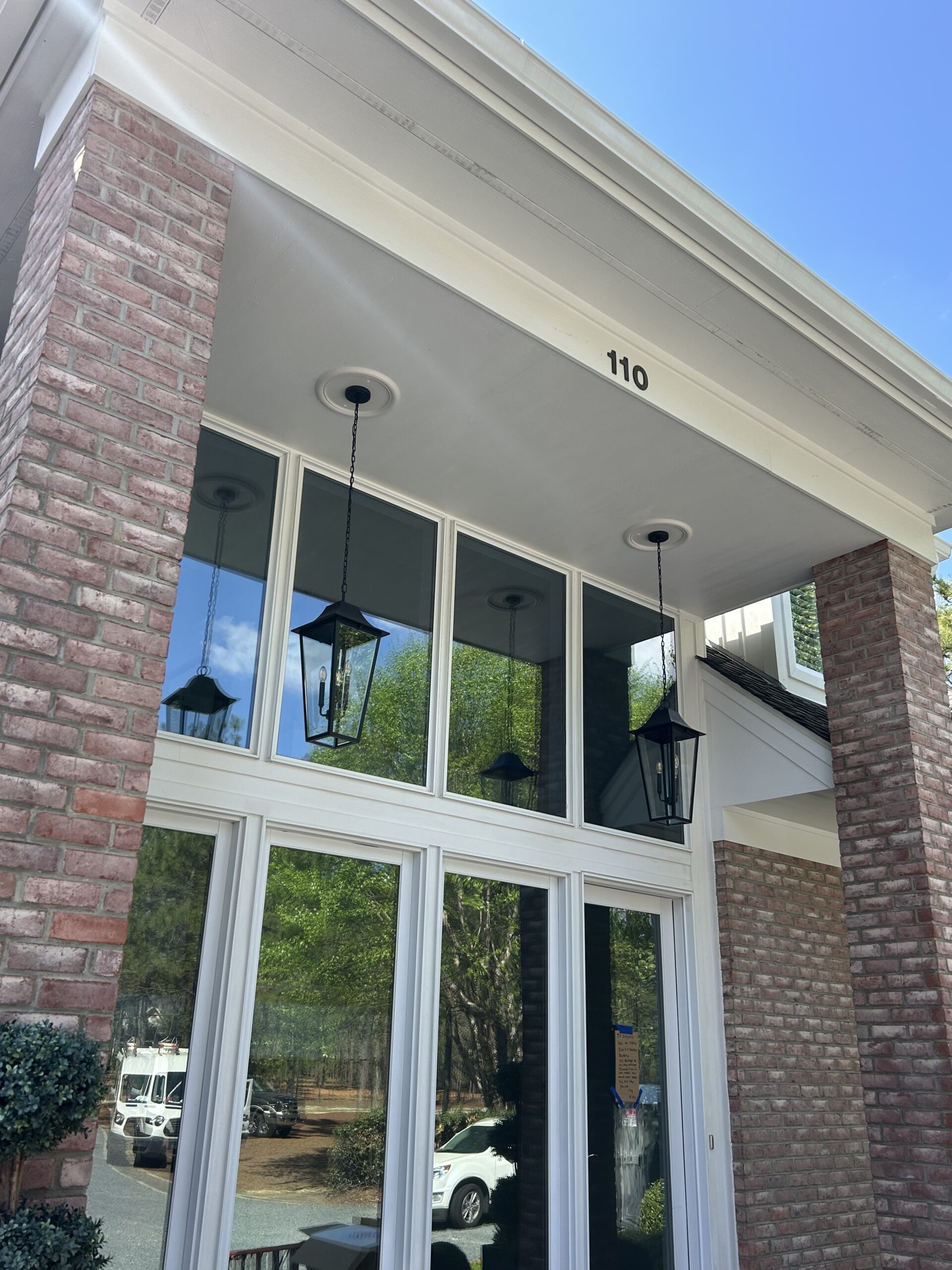 Brick building entrance with large glass windows, two black lantern-style hanging lights, and the number 110 above the doorway. Trees and parked vehicles are visible in the reflection.